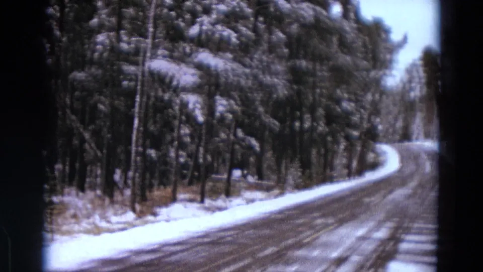 A winding snow-covered Michigan road curves through dense winter trees in 1967 8mm home-movie footage.