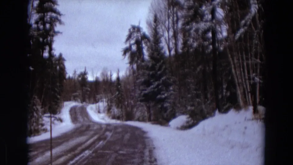The road rises and curves through a snow-covered wooded area, filmed from a moving vehicle on 8mm film.