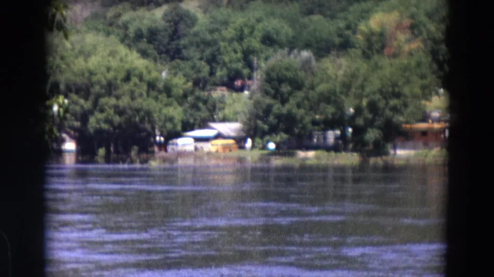 1967 home-movie still showing rippled lake water, dense green trees, and distant pale boats or structures along the bank.