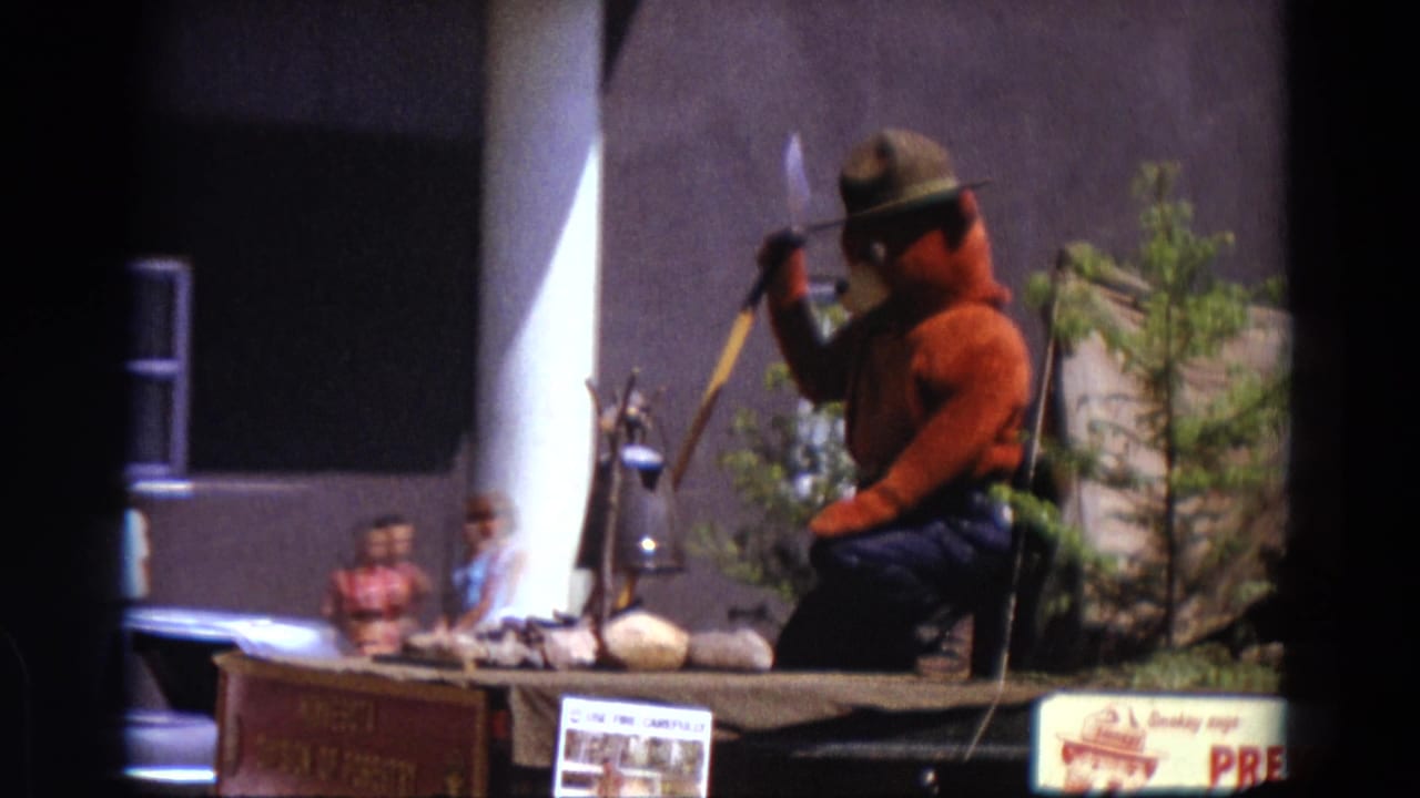 Smokey Bear figure on a parade float with shovel, tent display, signage, and spectators nearby.