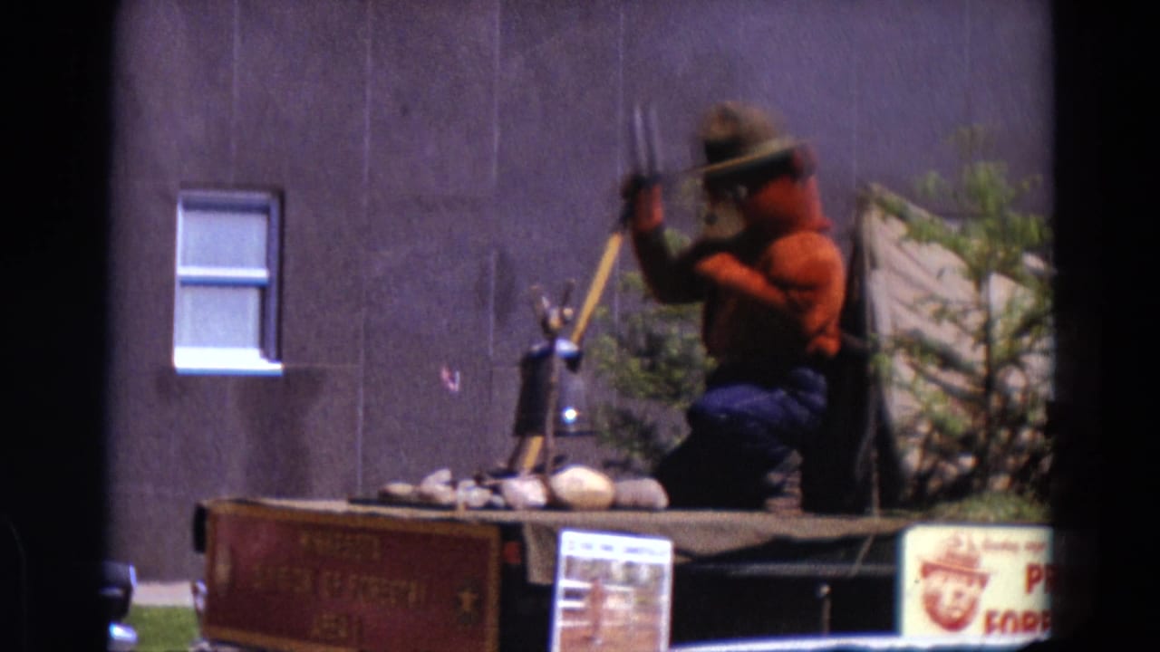 Smokey Bear float seen closer with shovel, small tent, rocks, plants, and a windowed wall behind.