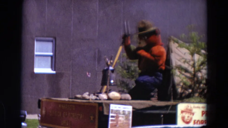 Smokey Bear stands on a Detroit parade float holding a shovel beside tent and rock props, with plants and a building wall behind.
