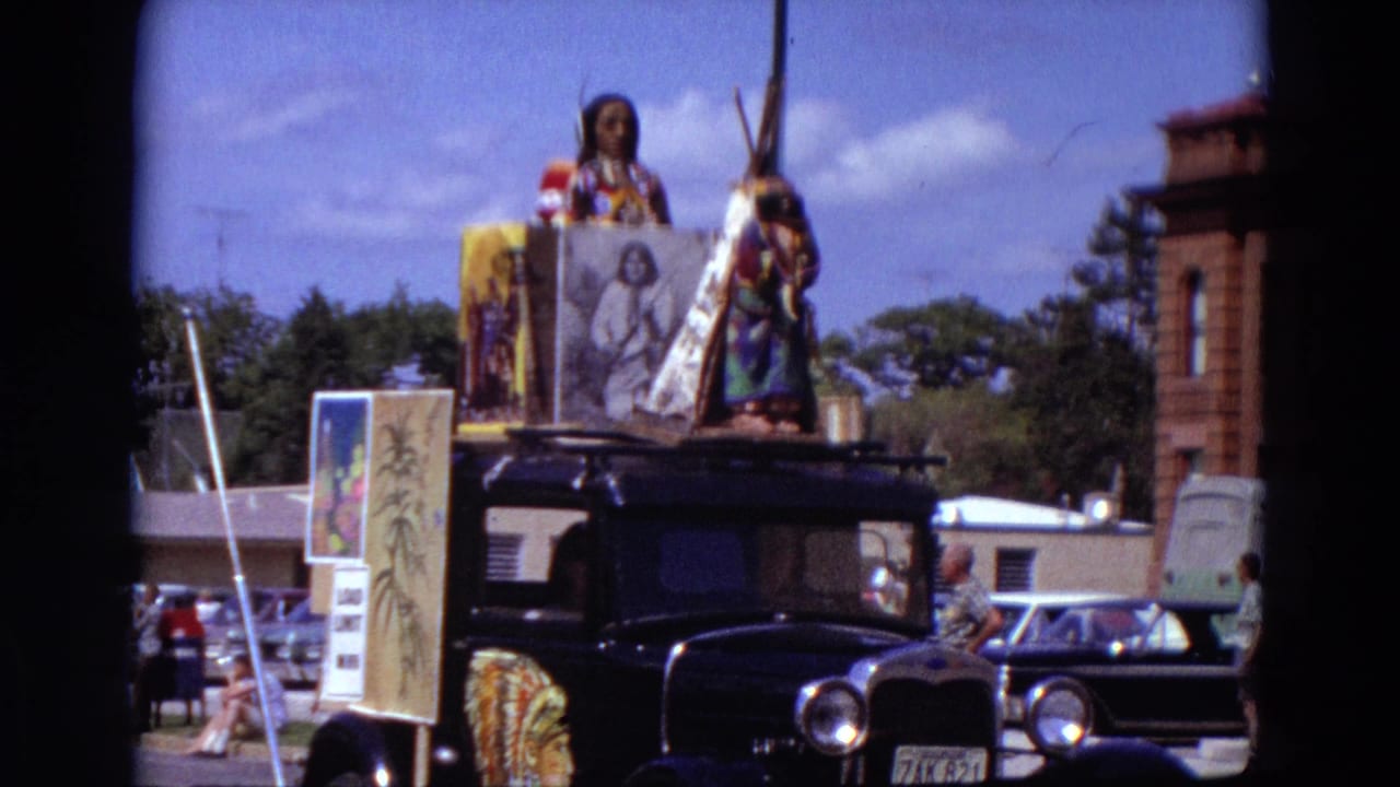 1967 Detroit parade frame showing a black vintage car decorated with Native American-themed figures, a teepee, signs, and street traffic behind.