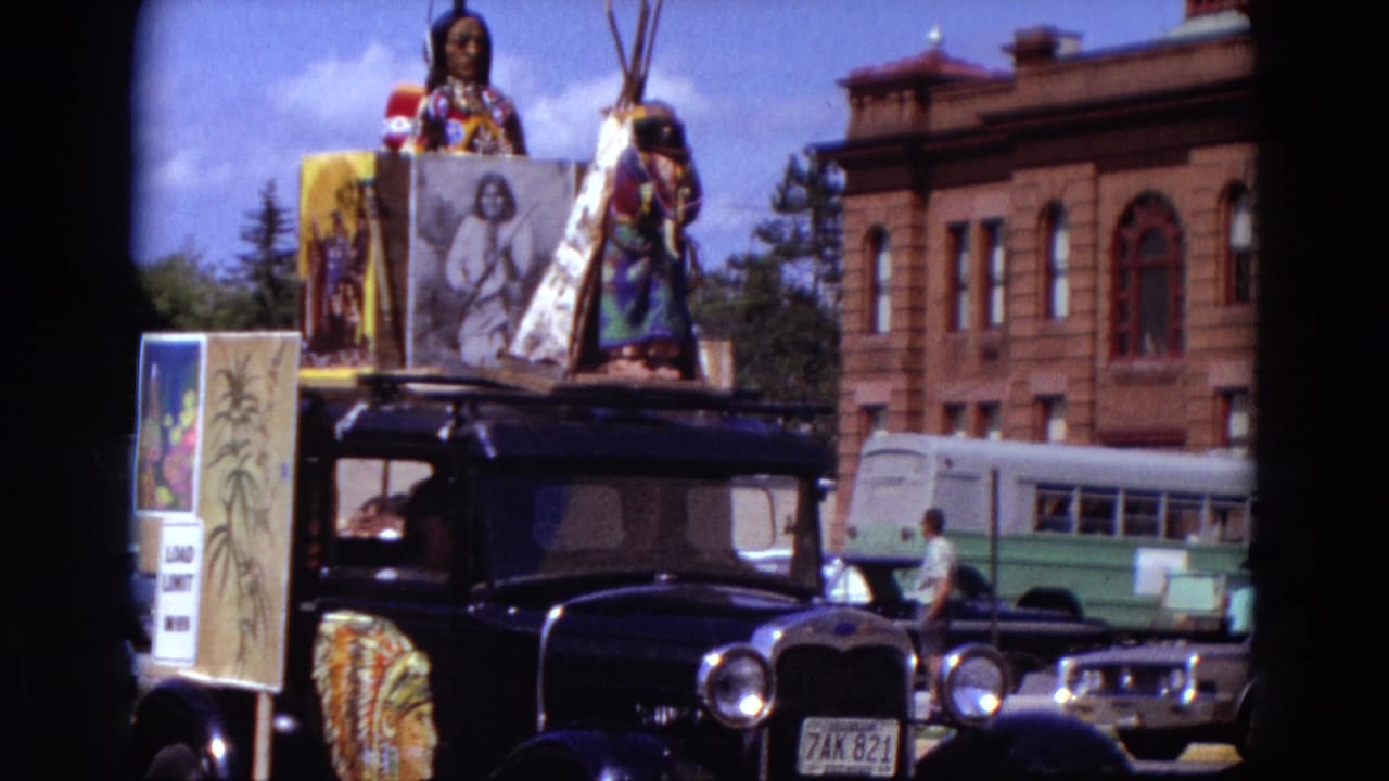 A black parade car in 1967 Detroit carries Native American-themed decorations and a teepee prop, with buses, cars, and a brick building in the background.