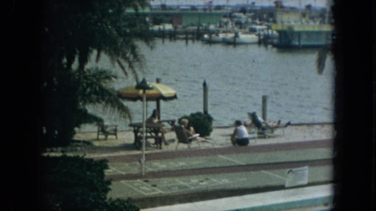 1967 Florida 8mm frame of people relaxing beside a waterfront walkway, with palm trees, a striped umbrella, lounge chairs, and marina boats beyond.