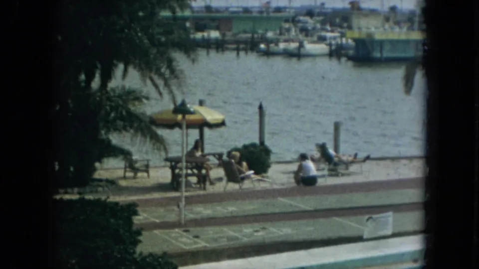 1967 Florida 8mm frame of people relaxing beside a waterfront walkway, with palm trees, a striped umbrella, lounge chairs, and marina boats beyond.