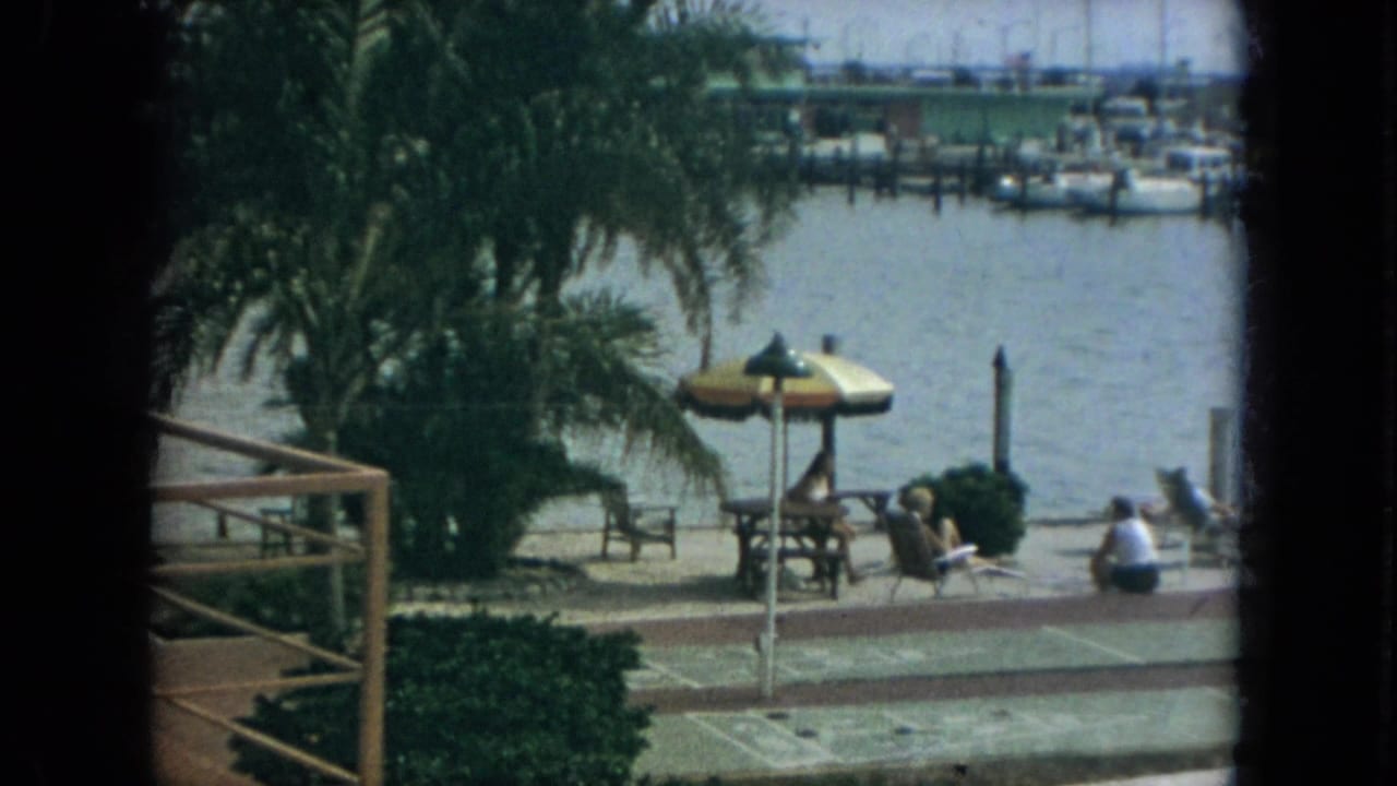 1967 Florida 8mm still showing a marina-side walkway, palm fronds, umbrella shade, seated people, lounge chairs, and a railing at the edge of frame.