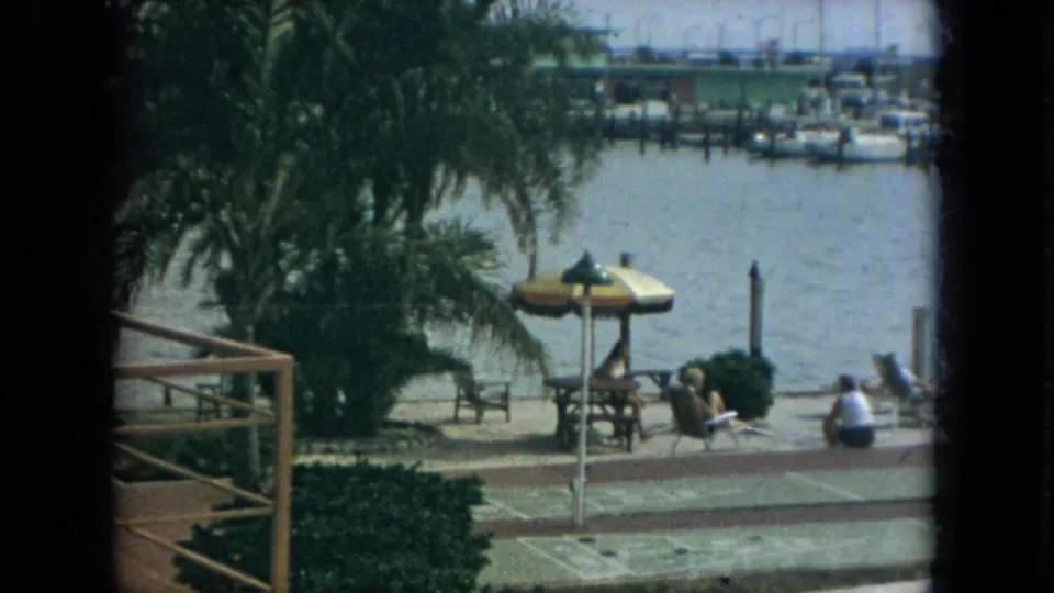 1967 Florida 8mm still showing a marina-side walkway, palm fronds, umbrella shade, seated people, lounge chairs, and a railing at the edge of frame.