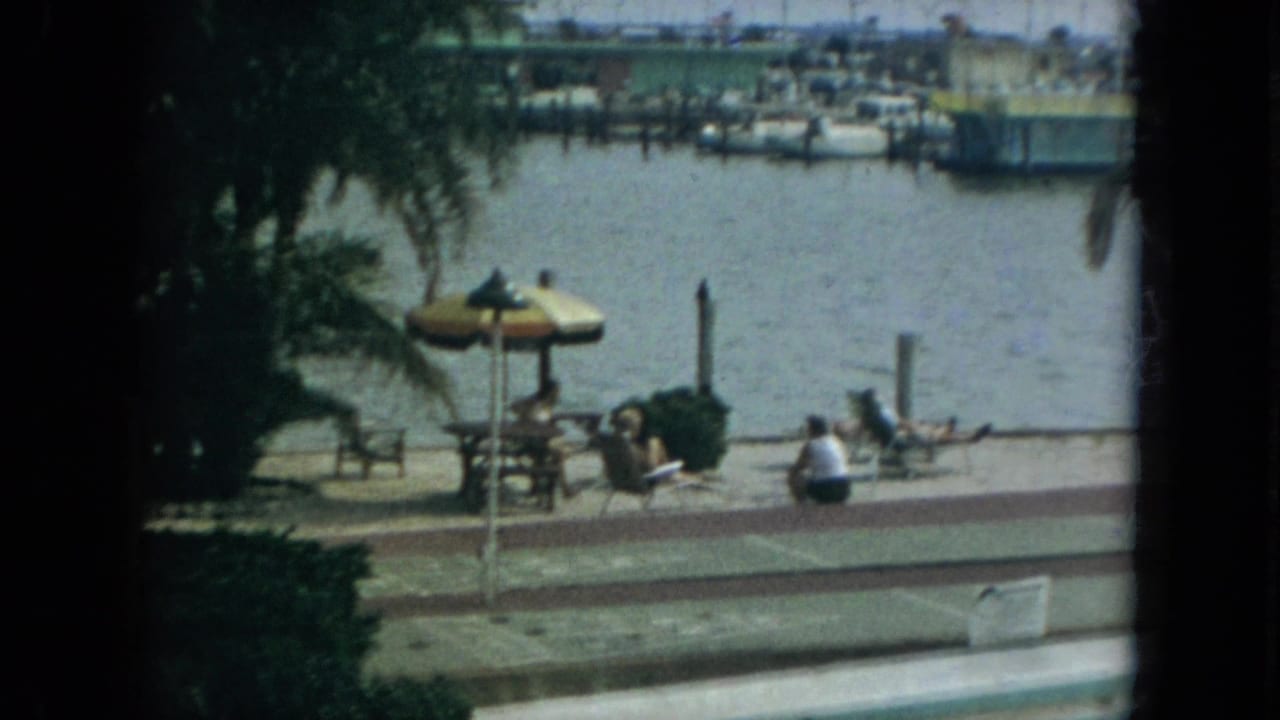 1967 Florida home-movie frame of people relaxing by a waterside walkway, with lounge chairs, a striped umbrella, palm trees, and marina boats in view.