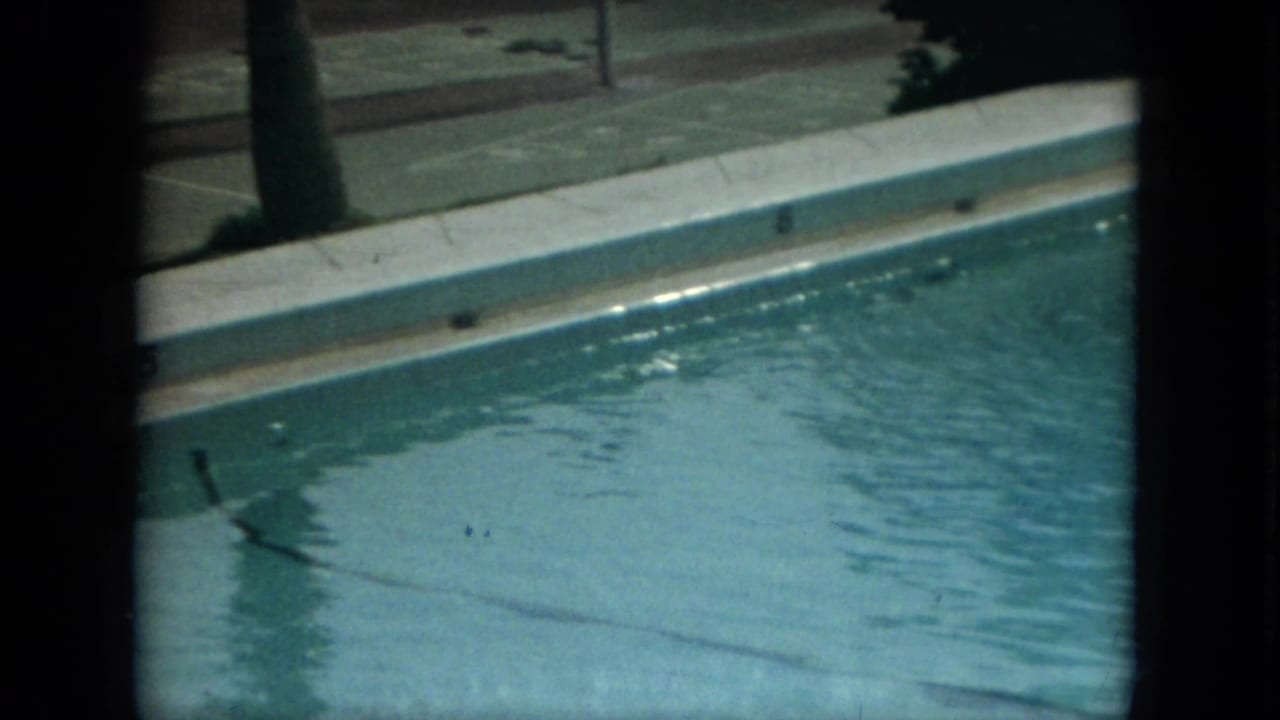 1967 Florida home-movie frame showing an empty resort swimming pool with pale deck edges, water ripples, and metal handrails.