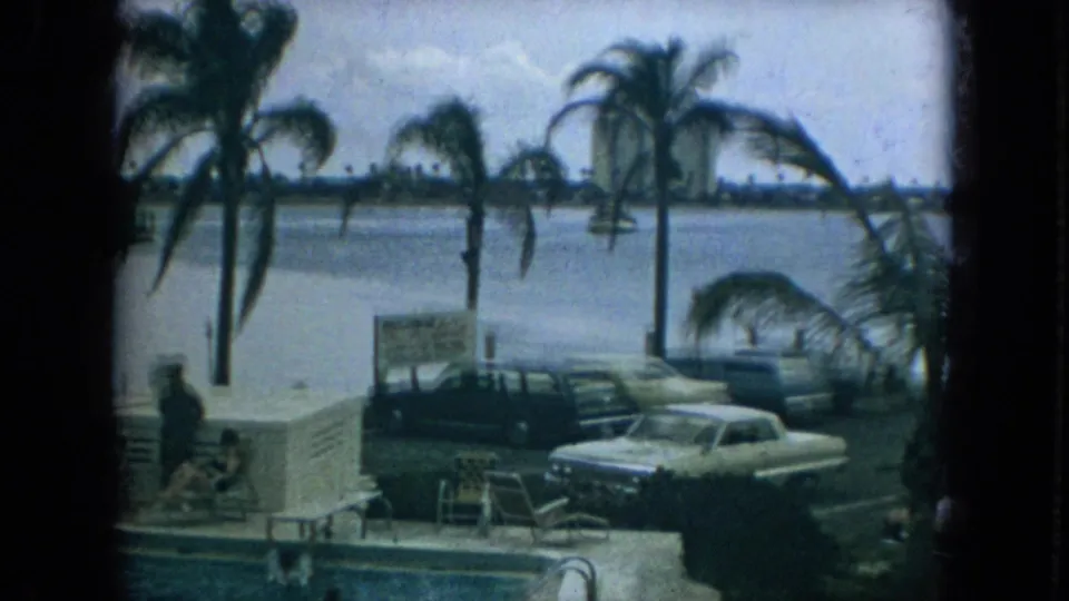 1967 Florida home-movie view of palm trees by the water, parked classic cars, a visible sign, pool steps, and distant shoreline buildings.