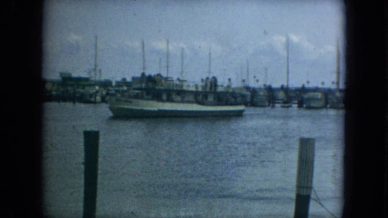 1967 Florida harbor view with a large boat moving past dock pilings, marina structures, and a line of sailboat masts.