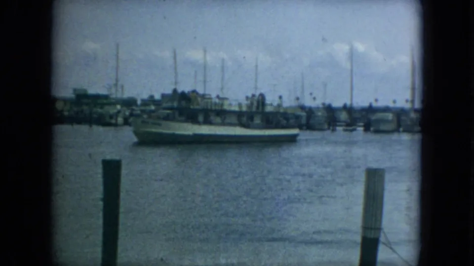 1967 Florida harbor view with a large boat moving past dock pilings, marina structures, and a line of sailboat masts.