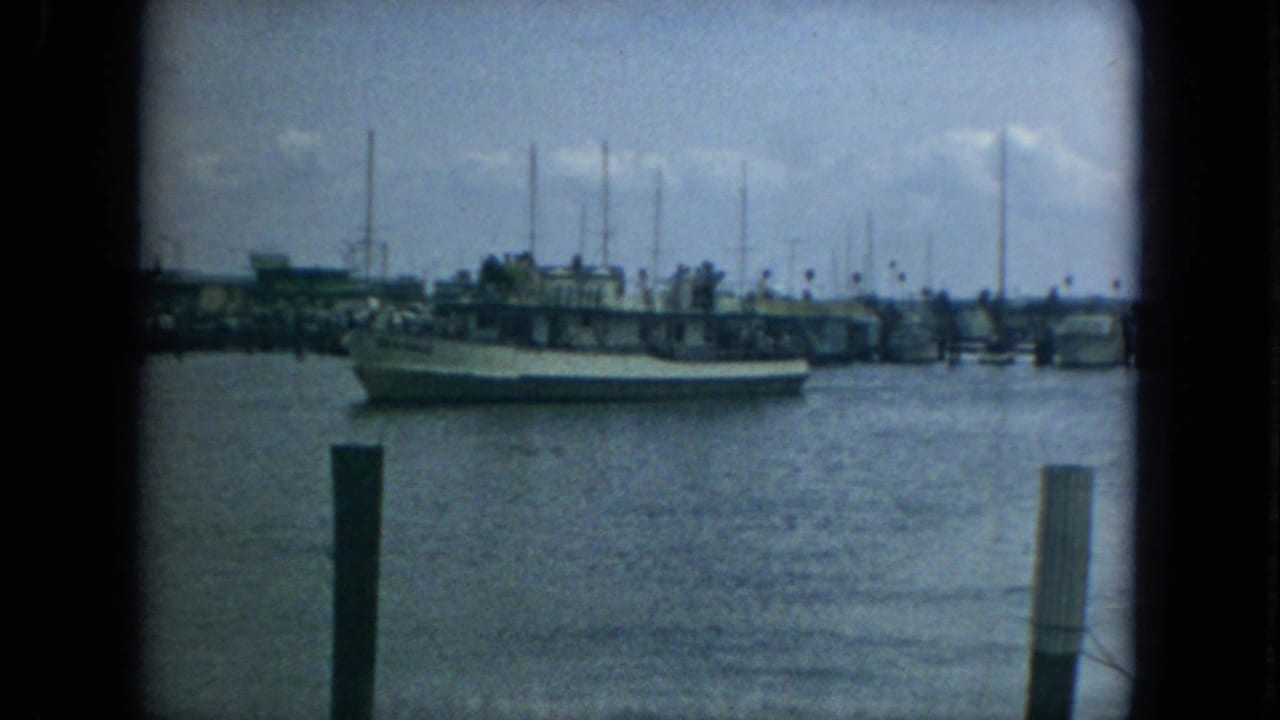 1967 Florida 8mm frame of a boat crossing calm harbor water with dock pilings, marina buildings, and sailboat masts behind it.