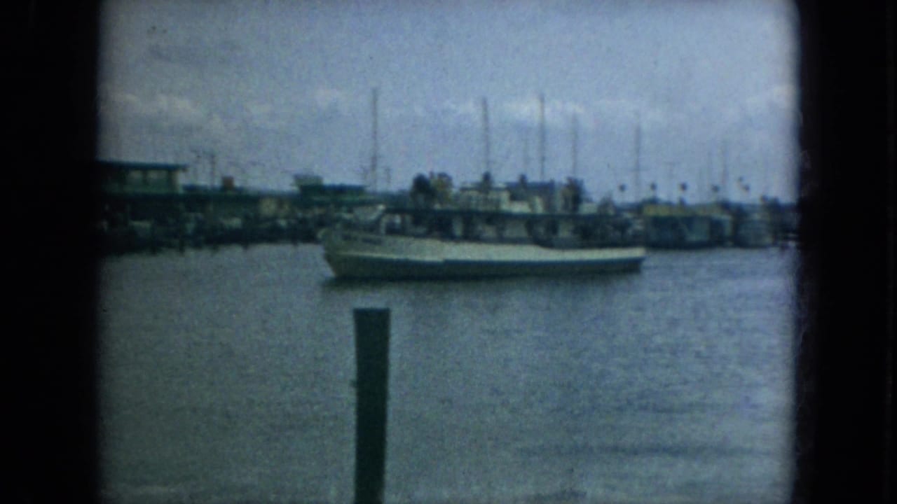1967 Florida harbor frame showing a large boat near dock pilings with sailboat masts, waterfront structures, and calm water beyond.