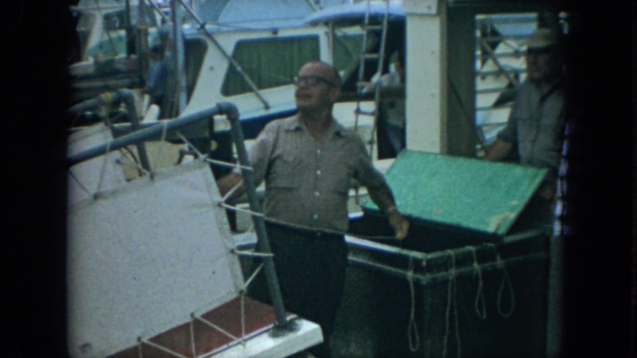 In 1967 Florida home-movie footage, an older man in glasses stands near a green cooler on a boat deck with marina boats behind him.