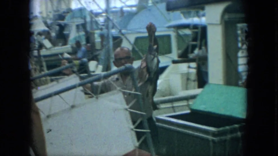 The man raises a fish on a stringer on a Florida marina boat deck, with a green cooler and nearby vessels visible behind him.