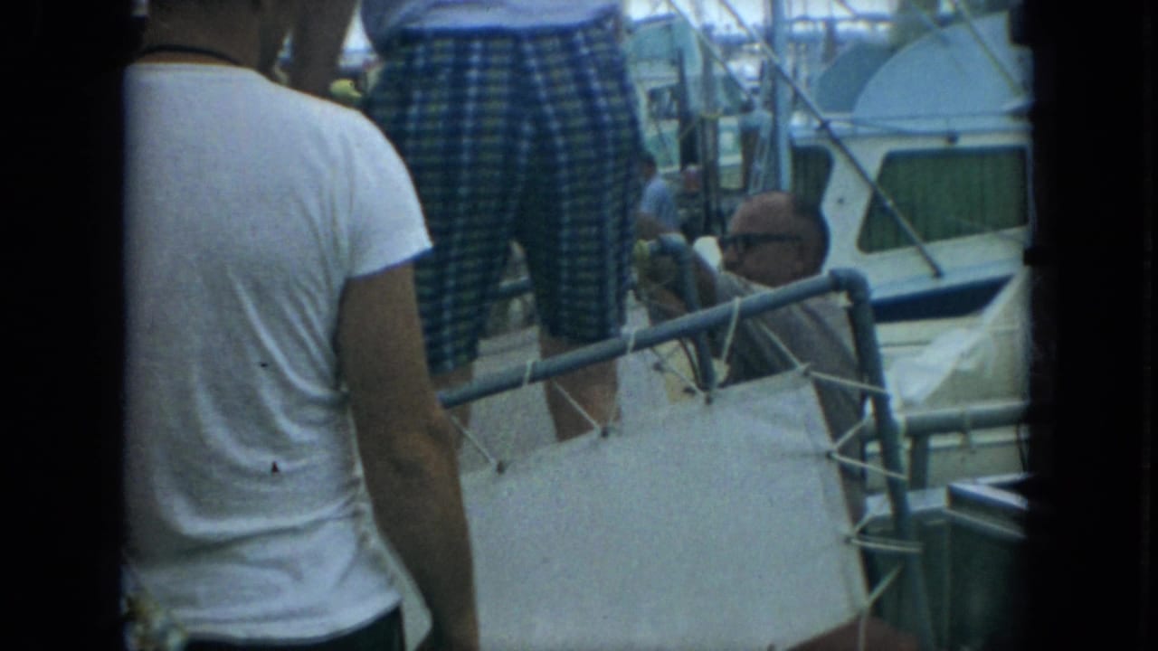 Foreground people partly obscure the older man near the boat railing, with cabin boats and marina activity in the background.