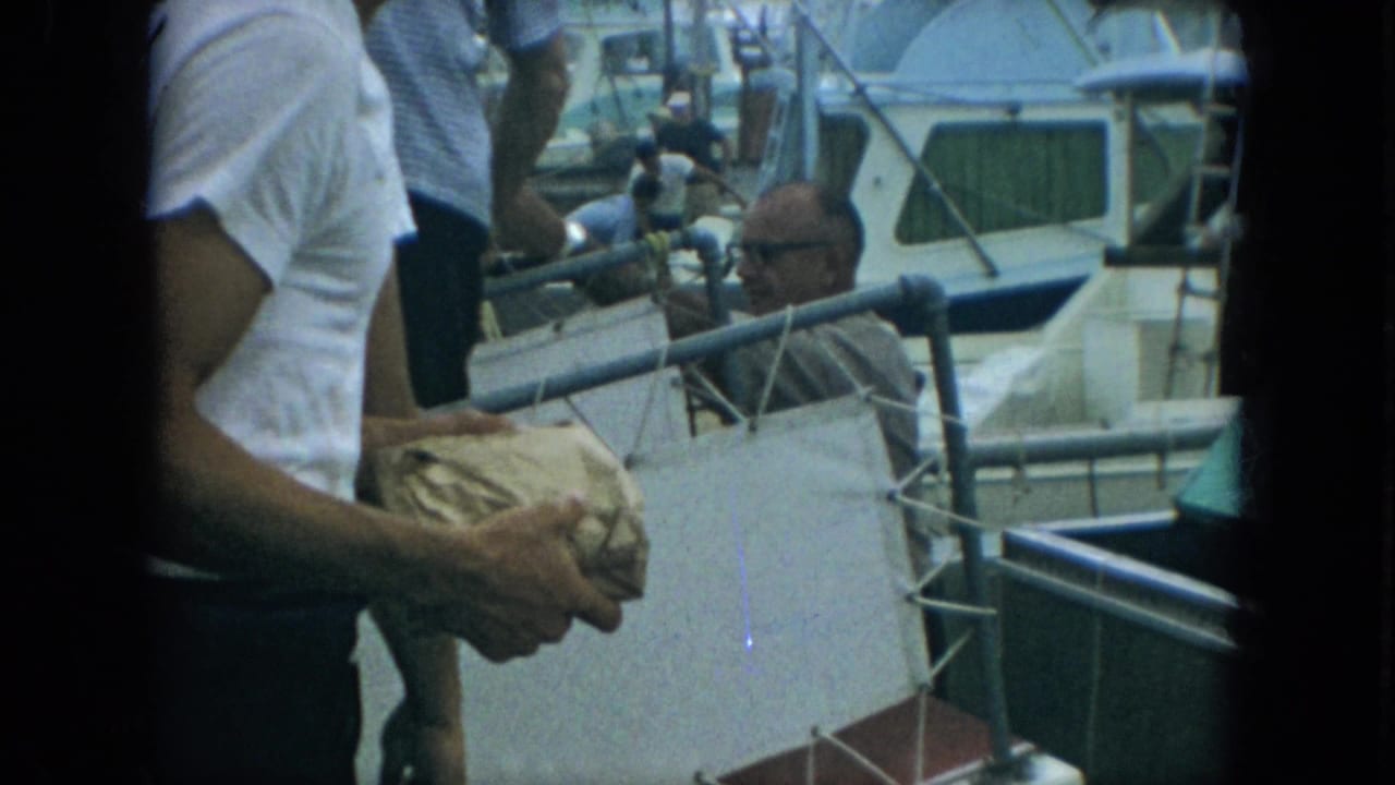 1967 Florida 8mm frame of a man in a white shirt holding a wrapped fish near a marina dock and small boats.