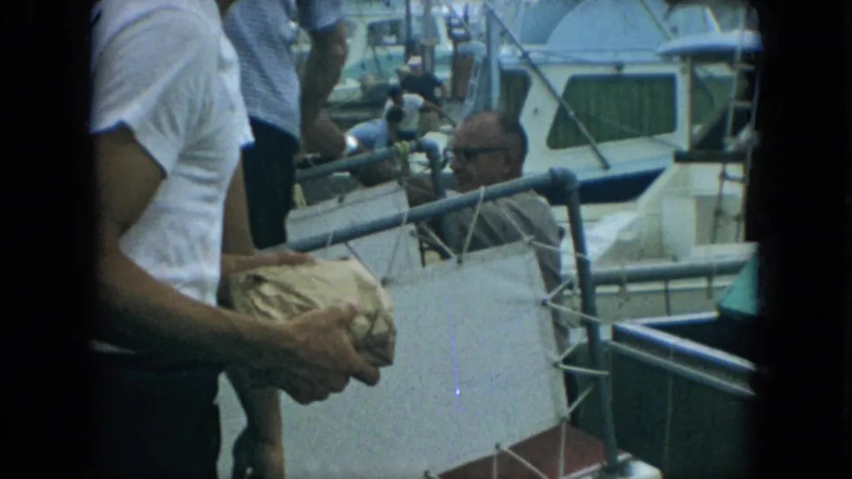 1967 Florida 8mm frame of a man in a white shirt holding a wrapped fish near a marina dock and small boats.