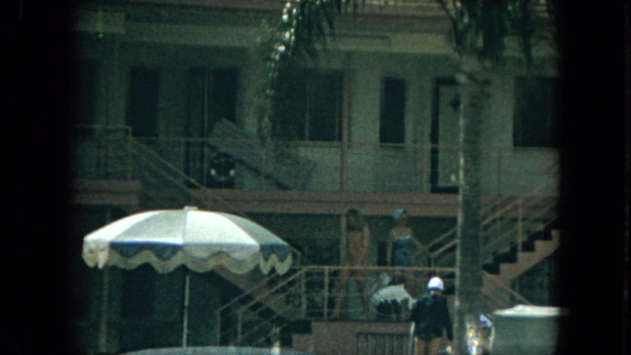 Two-story motel exterior with stairs, palm fronds, guests, and a striped patio umbrella.