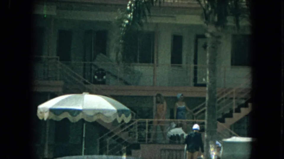 1967 Florida motel exterior with outdoor stairs, palm fronds, distant guests, and a striped patio umbrella in the foreground.