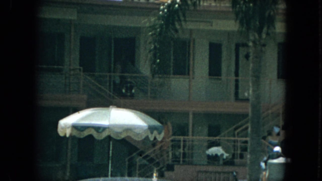 1967 Florida home-movie frame of a motel exterior with striped umbrella, railings, stairs, palm fronds, and distant guests near the walkway.