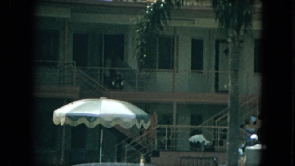 1967 Florida home-movie frame of a motel exterior with striped umbrella, railings, stairs, palm fronds, and distant guests near the walkway.