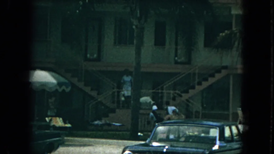 1967 Florida apartment scene with a woman moving down exterior stairs above parked mid-century cars.