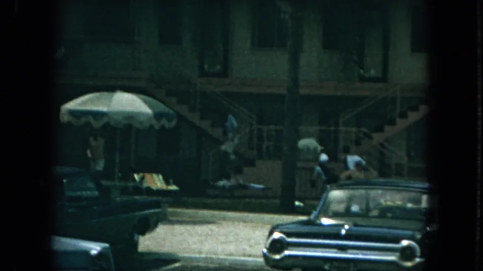 1967 Florida apartment courtyard with a woman on exterior stairs, palm trees, a patio umbrella, and vintage cars.