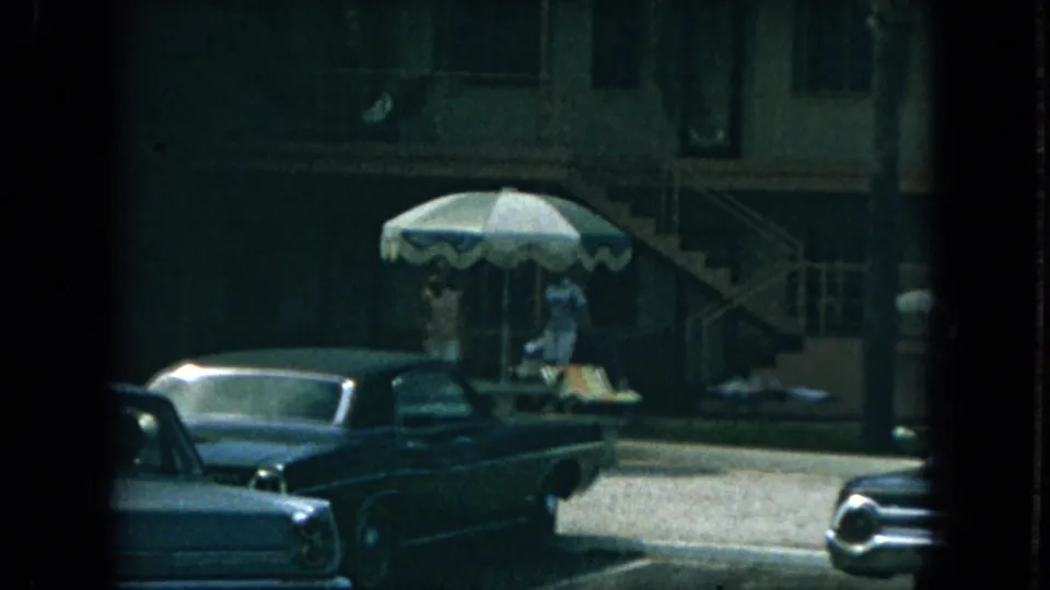 1967 Florida apartment exterior showing people near a striped patio umbrella, exterior stairs, palm trees, and parked cars.