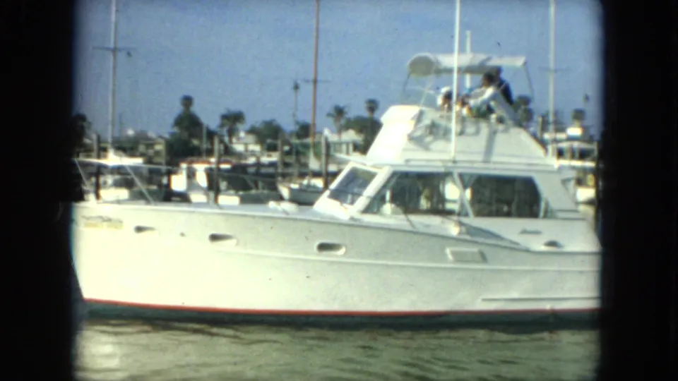 1967 Florida home-movie frame showing a white cabin boat at a marina with distant people and other vessels in the background.