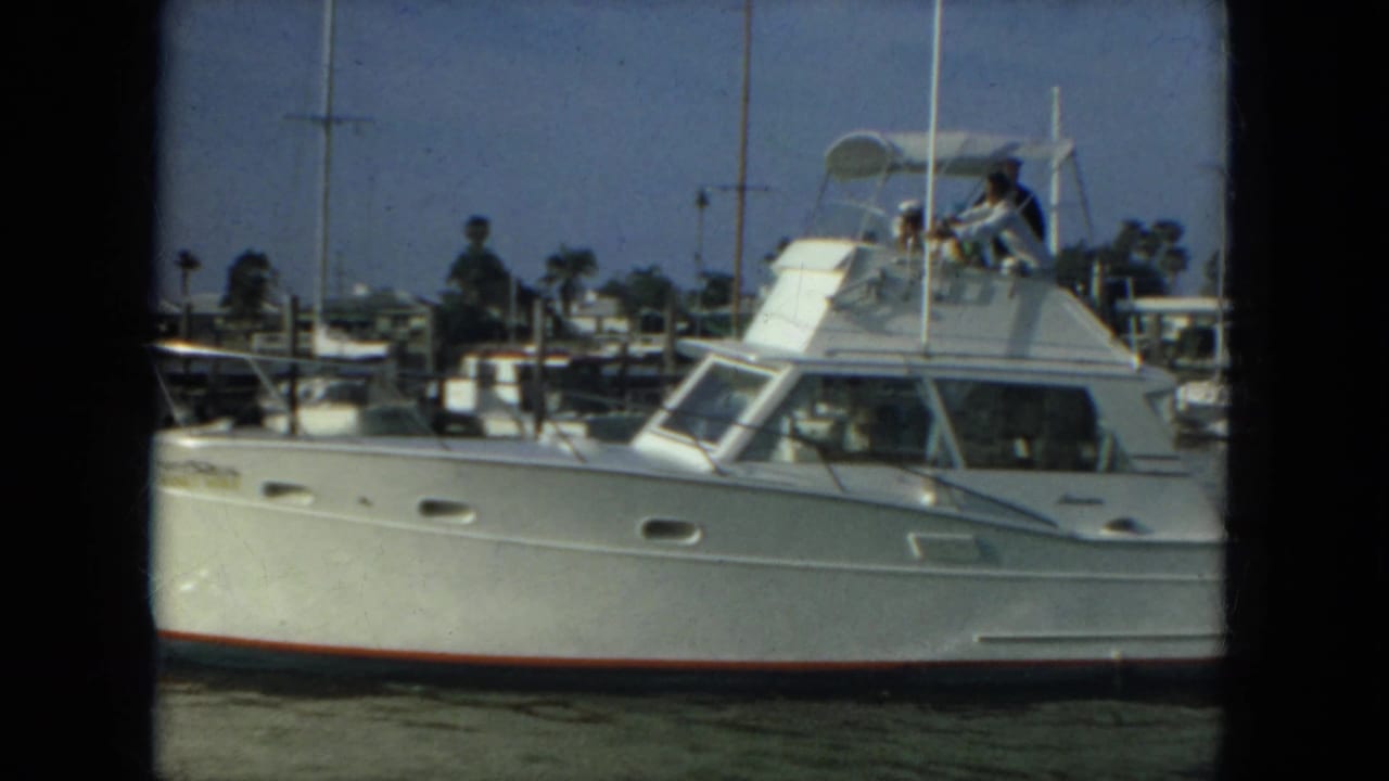 1967 Florida 8mm frame of a white motor yacht moving through a calm marina with passengers visible on the upper deck.