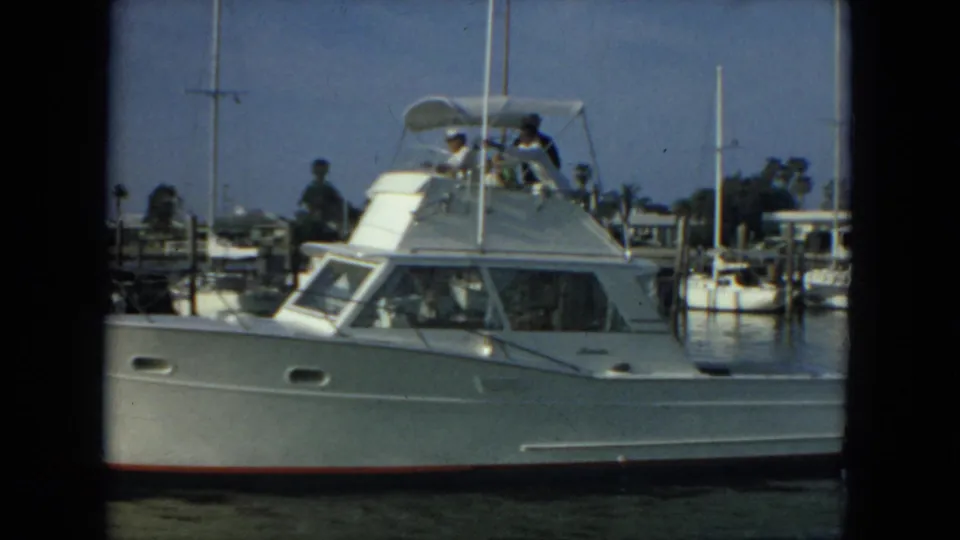 1967 Florida harbor view with a white yacht, upper-deck passengers, sailboat masts, docks, and palm trees in the background.