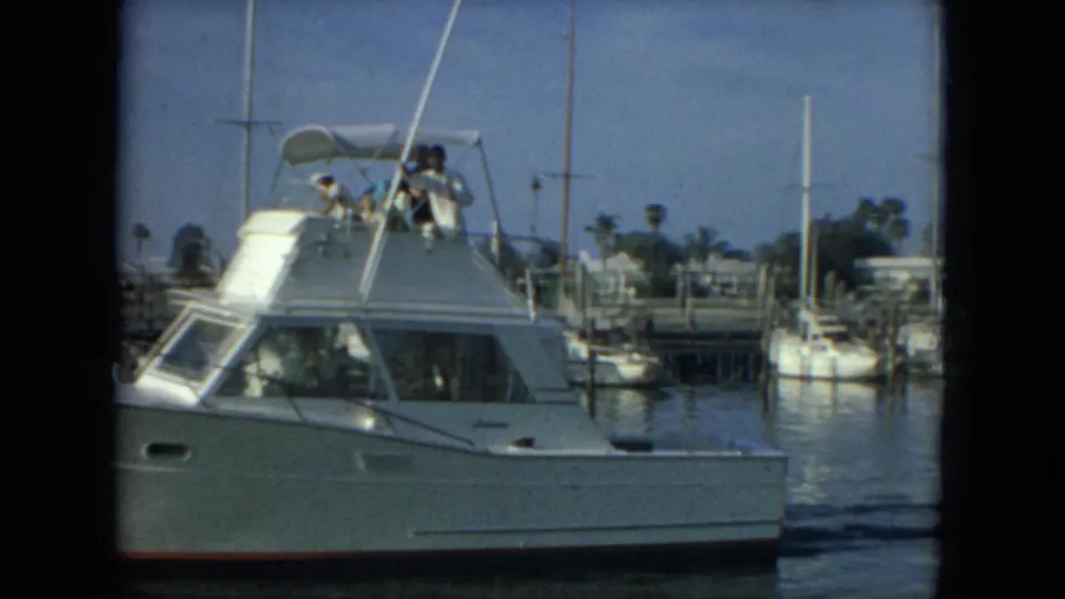 White motor yacht partly framed in close view as it passes through a Florida marina, with passengers and docked boats behind it.