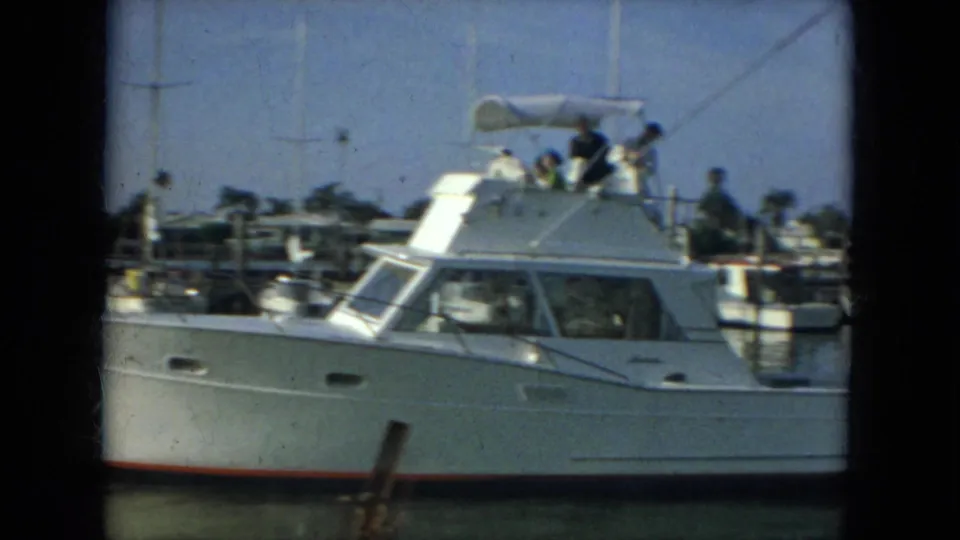 1967 Florida home-movie frame of a white motor yacht in profile with people on the flybridge and marina docks behind it.