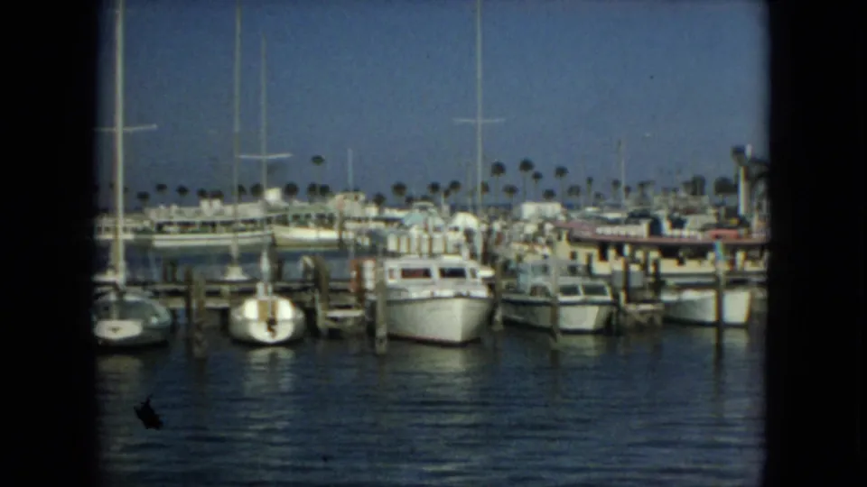 1967 Florida 8mm frame showing a broad marina view with moored boats, wooden piers, sailboat masts, and palm trees in the background.