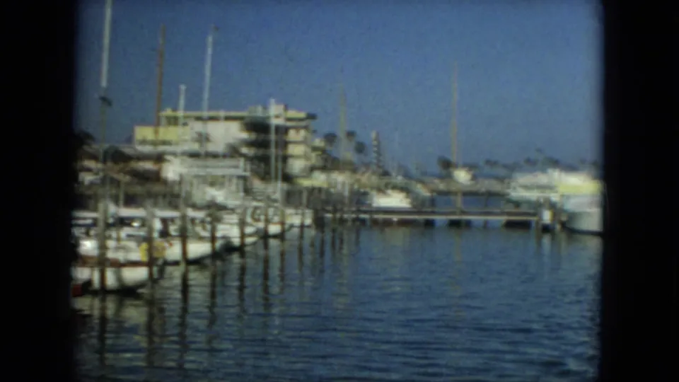 1967 Florida waterfront view with docked boats, wood pilings, sailboat masts, a low marina building, and blue water under clear sky.
