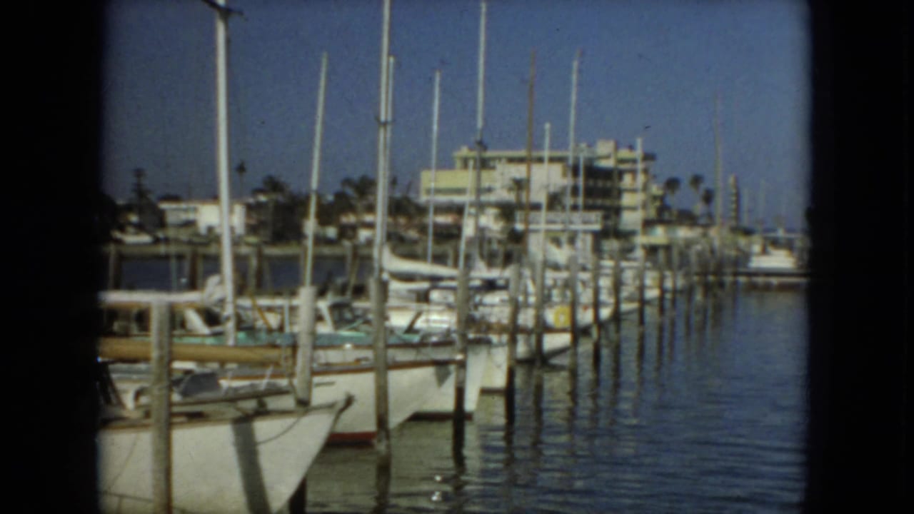 1967 Florida 8mm frame showing moored sailboats, dock pilings, masts, calm marina water, and low waterfront buildings.