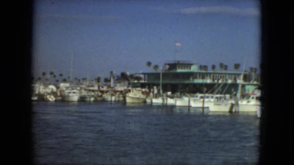 1967 Florida marina view with yachts, wooden docks, palm trees, a green-roofed waterfront building, and a small American flag.