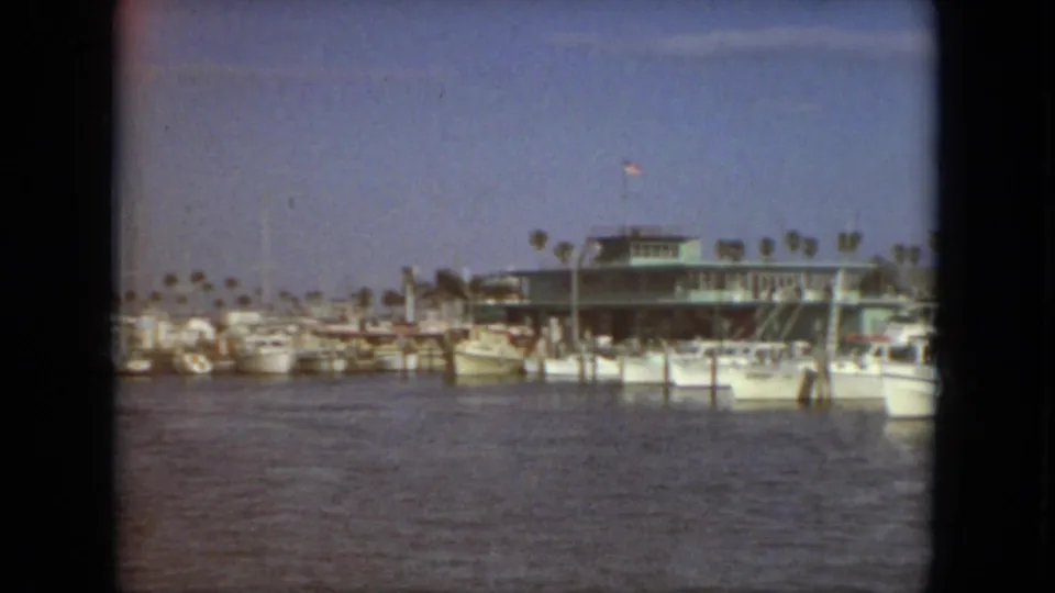 1967 Florida 8mm frame of a marina basin with moored boats, dock posts, palm trees, and a low waterfront building.