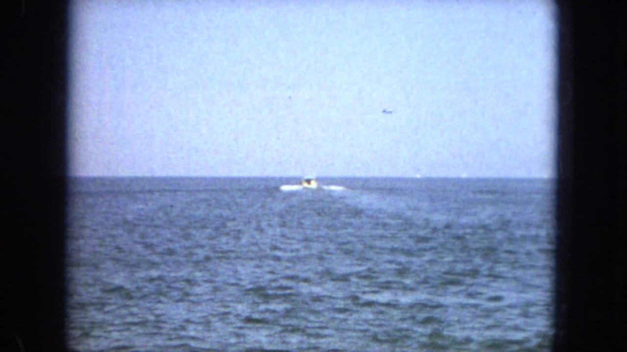 Small boat crossing distant blue water with a white wake under pale sky.
