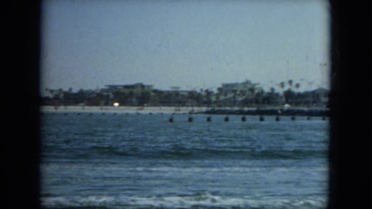 Distant shoreline across blue water with surf, palm trees, and low buildings.