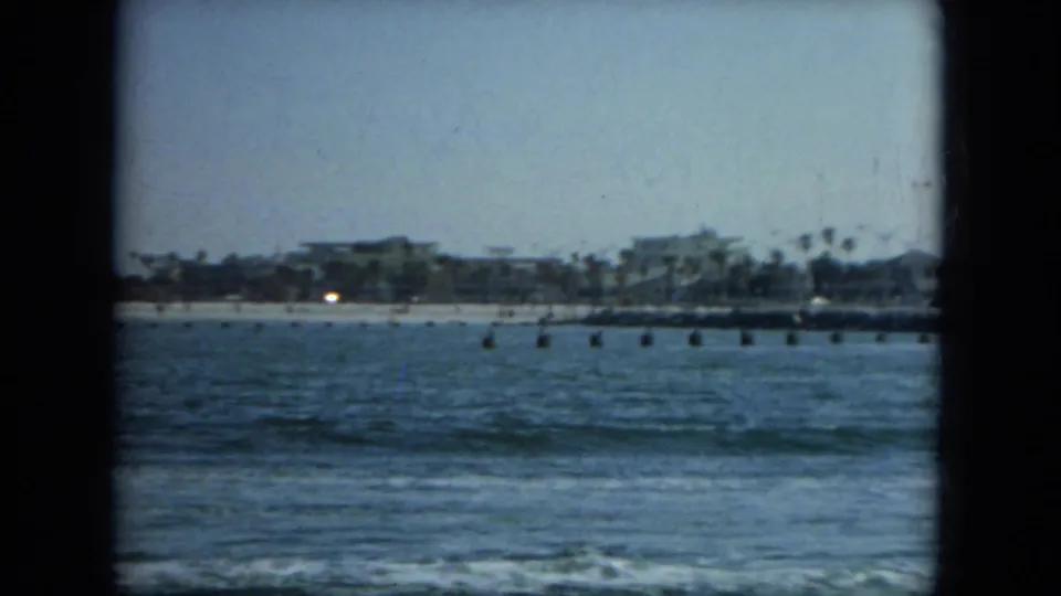 1967 Florida coastal view across blue water, with gentle surf, a sandy shore, palm trees, and low beachfront buildings in soft 8mm focus.