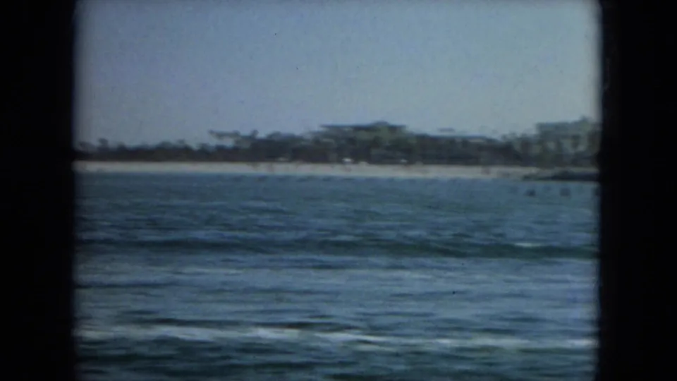 1967 Florida shoreline view with open water in the foreground and distant low buildings behind a pale beach under a hazy sky.