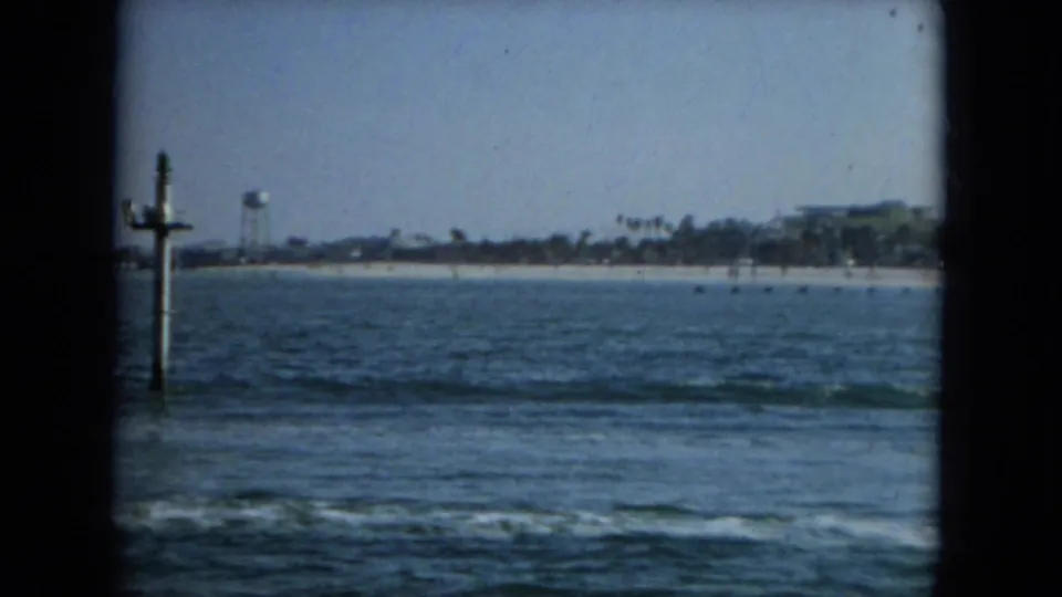 1967 Florida waterfront frame showing blue water, low surf, a sandy beach, a water tower, palm trees, and distant shoreline buildings.