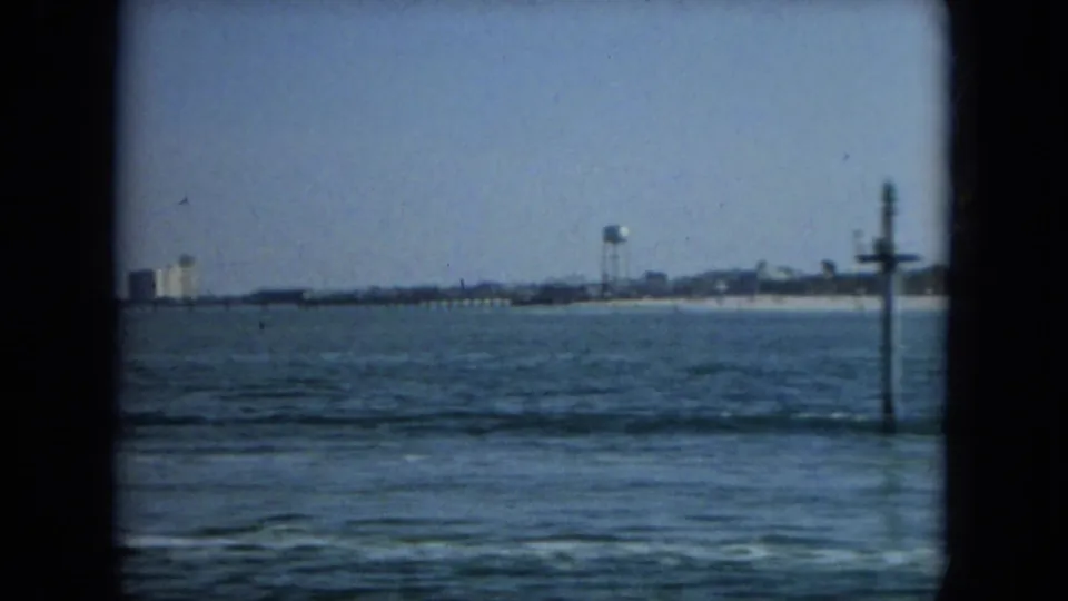 1967 Florida coastal view with waves in the foreground, a distant beach, a water tower, industrial-looking structures, and a shoreline post.