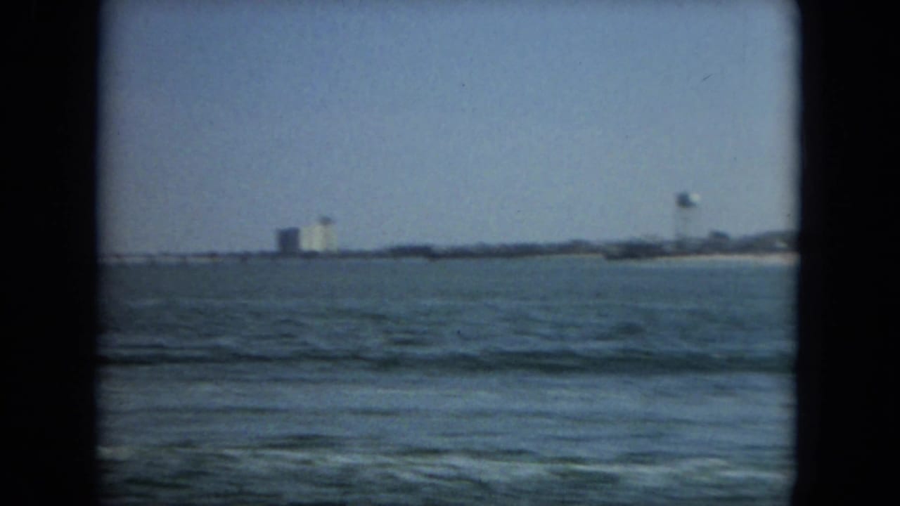 1967 Florida 8mm frame showing open sea in the foreground and a distant beach town with a water tower on the horizon.
