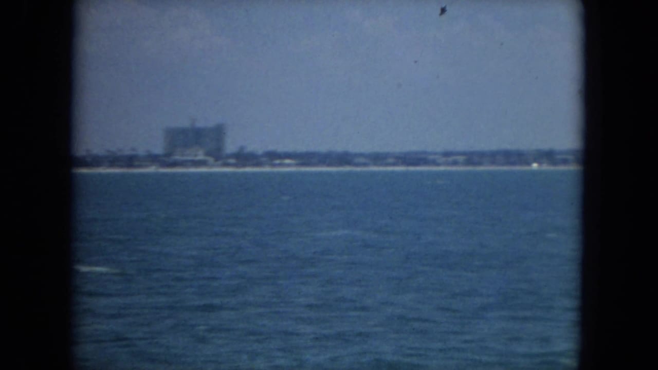 Open water fills the foreground with a distant beach and blocky buildings on the horizon.