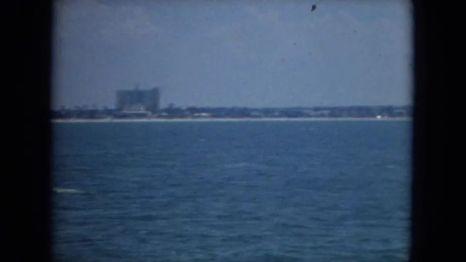 1967 Florida coastal view from the water, with a pale shoreline and distant low-rise buildings in soft focus.